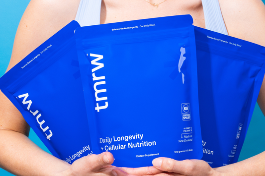 Woman holding three blue Tmrw Daily Longevity + Cellular Nutrition supplement pouches against a blue background.